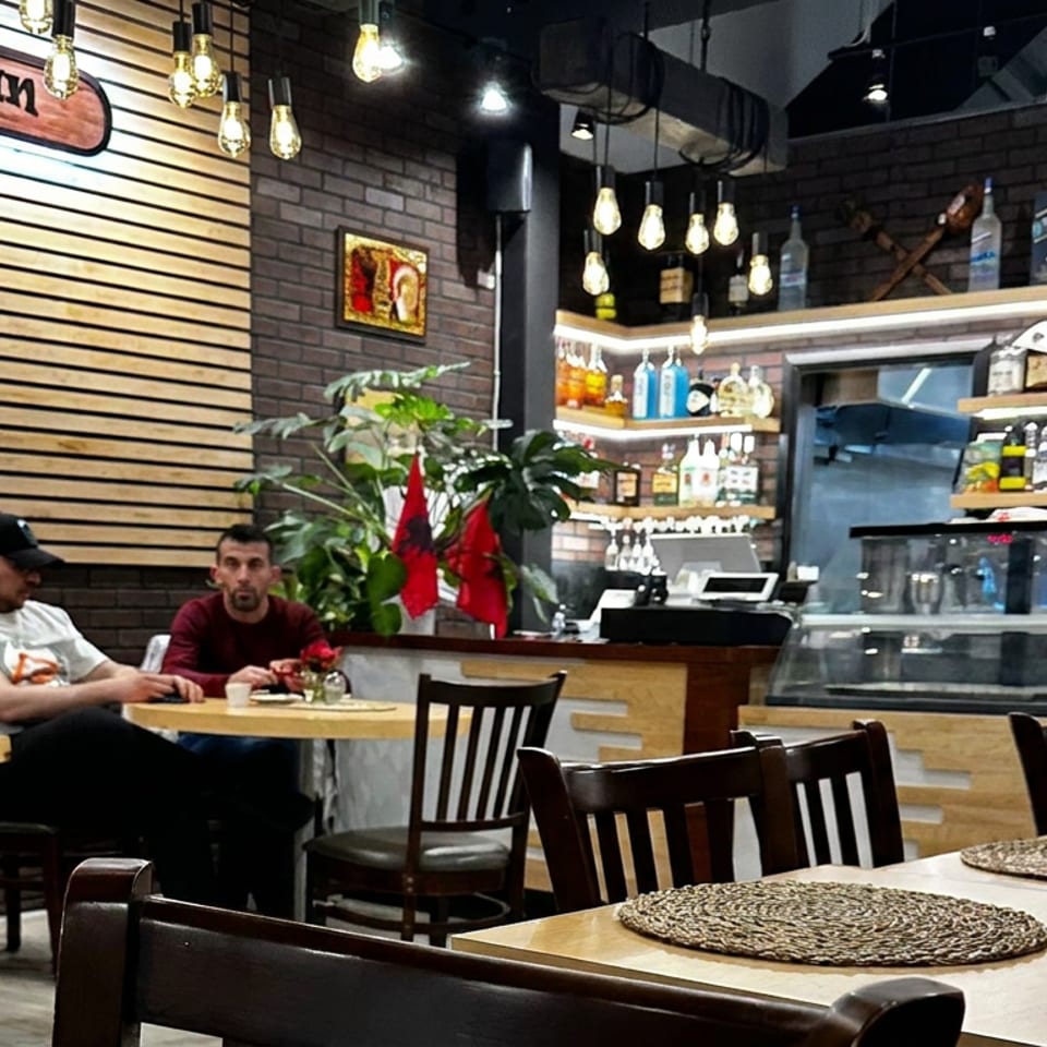 Ballkan Grill dining room with two diners, wooden tables, pendant lights, and a stocked bar beneath brick walls.