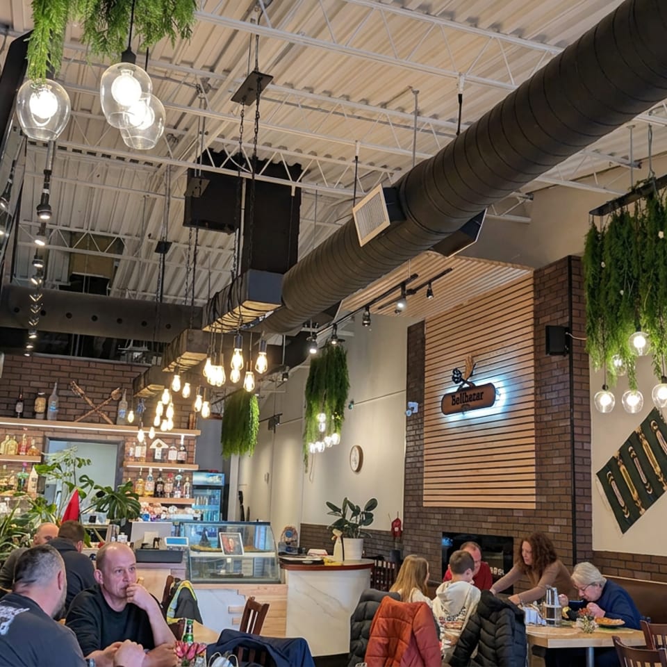 Industrial-style restaurant interior with brick walls, hanging plants, pendant lights, bar counter, and diners seated at tables.