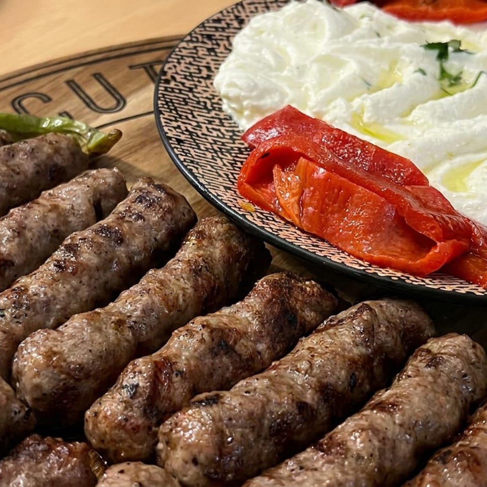 Grilled sausages served with creamy salad, sliced tomatoes, olives, and green peppers on a wooden table.