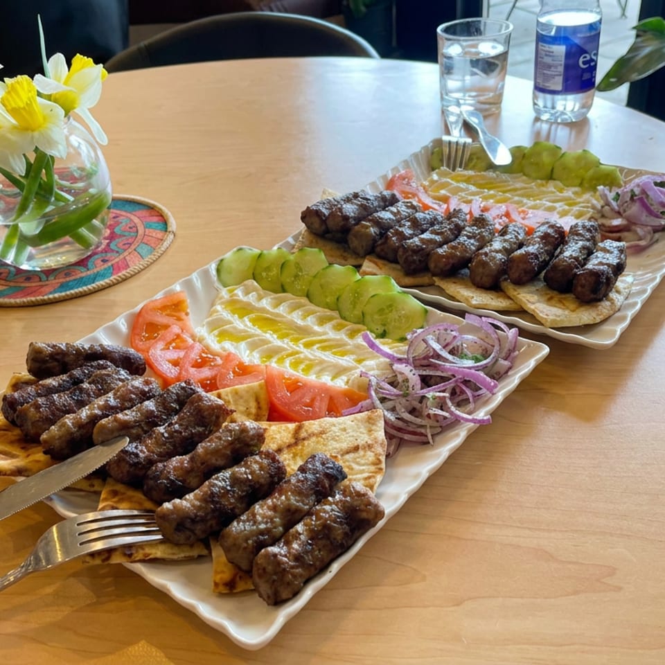 Two platters of grilled minced meat sausages with flatbread, sliced tomatoes, cucumbers, and onions on a restaurant table.