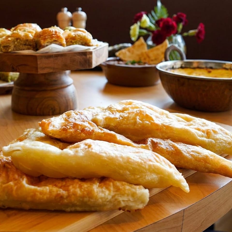 Golden fried pastries arranged on a wooden platter, surrounded by savory dishes, bread rolls, flowers, and dining table decor.